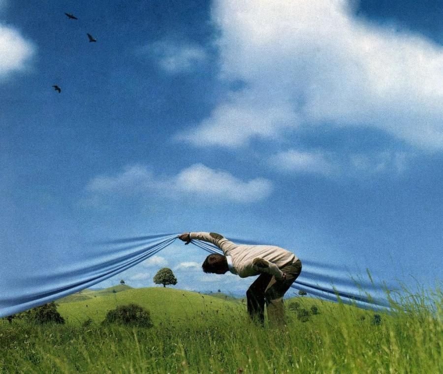 A person lifting a curtain of blue sky to reveal a green landscape.